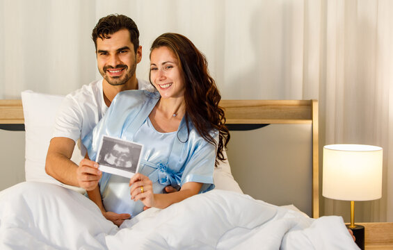 Cheerful Caucasian husband tenderly embrace lovely pregnant wife on bed under quilt and smile to show x-ray film image of expecting unborn baby