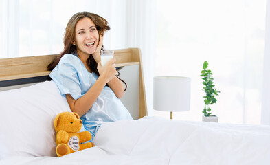 Cheerful Caucasian pregnant woman smiling as enjoy drinking glass of milk for health nourishment to prepare birth of unborn baby while resting on bed beside teddy bear in bedroom