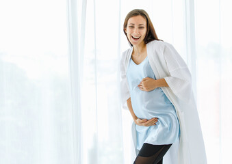 Lovely pregnant woman on maternity dress and nightgown tenderly embracing belly of unborn baby with care and smile as happy to become motherhood