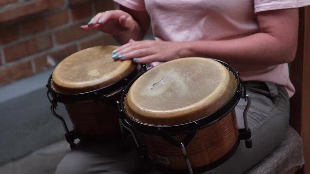 Girl in pink shirt sits on chair, holds mini bongos on her knees and plays on them. Close up of the hands of girl musician with manicure playing traditional ethnic percussion instrument bongo drums