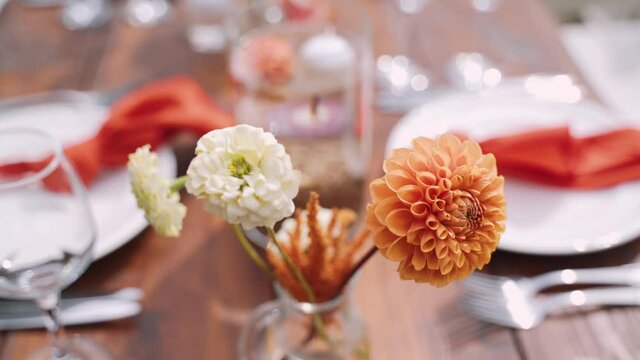 Orange, coral and beige zinnia flowers stand in a vase on a banquet table, close-up. Festive wooden table decor with plates, cutlery and glasses.