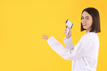 Female trainee doctor with thermometer gun on yellow background