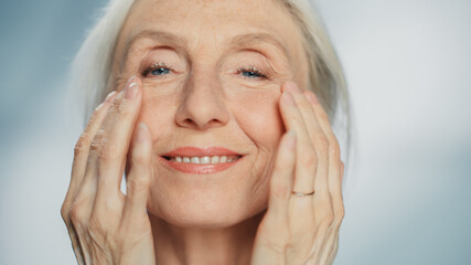Portrait of Senior Woman Looking at Camera, Touching Beautiful Face, Smiling. Elderly Female with Natural Grey Hair, Blue Eyes, Soft Skin. Dignified Old Age Concept for Skin care and Healthy Living