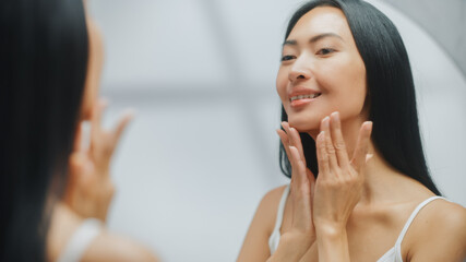 Portrait of Beautiful Asian Woman Gently Applying Face Cream Looking in Bathroom Mirror. Young Adult Female Makes Her Skin Soft, Smooth, Wrinkle Free with Natural Cosmetics Skincare Product