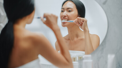 Portrait of Beautiful Asian Woman Uses Tooth Brush to Clean Her Natural White Teeth, Smiles in Bathroom Mirror Looking at Camera. Happy Female Doing Morning routine. Dental Hygiene, Wellness, Beauty