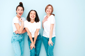 Fototapeta premium Three young beautiful smiling hipster female in trendy same summer white t-shirt and jeans clothes. Sexy carefree women posing near light blue wall in studio. Cheerful and positive models having fun