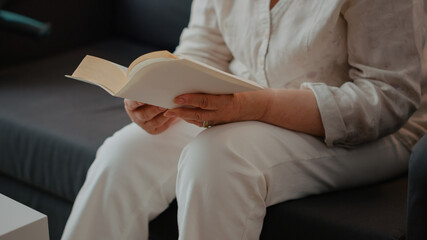 Grandmother reading novel story book in living room, enjoying retirement. Senior woman holding literature book to read for knowledge and intelligence, being concentrated and focused. Close up