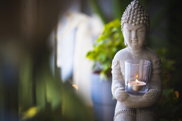 Buddha statue close-up in beautiful light. Close up of a Buddha figurine. Modern decor on a shelf, a buddha and a candle holder and a plants in a pot. Spiritual awakening - obrazy, fototapety, plakaty