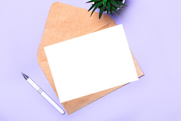 On a purple background there is a white pen, a potted plant, a craft envelope and a white blank card with a place to insert text. Top view. Template