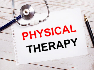 On a light wooden table there is a stethoscope, a pen and a sheet of paper with the text PHYSICAL THERAPY. Medical concept