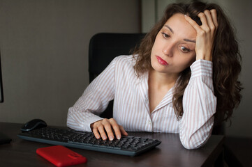 Tired young woman with curly hair and white shirt is working at the office using her laptop,...