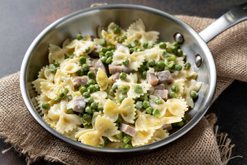 Farfalle with green peas, turkey ham and parmesan in cream sauce in a frying pan on the kitchen table. Traditional Italian pasta on a dark culinary background closeup