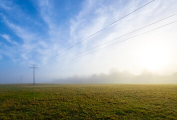 Overhead power line with wooden poles and electric wires spanning over rural landscape on a foggy and sunny winter morning near Tübingen Germany. Misty atmosphere scenery panorama after rainy night.