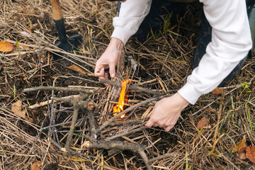 Close-up cropped shot of unrecognizable tourist man putting kindling on burning fire to keep warm and cook food at outdoors on overcast cold day. Concept of bushcraft, camping and survival in nature.