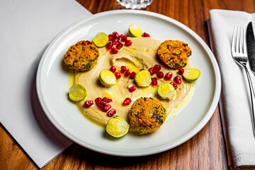 A close-up of falafel with humus, brussel sprouts and pomegranate seeds served on a white plate on a wooden table in the restaurant. A menu, fork and knife, napkin, glass of water.
