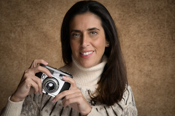 Retrato de joven mujer atractiva sonriente con cámara de fotos retro, con fondo desenfocado marrón y jersey blanco.