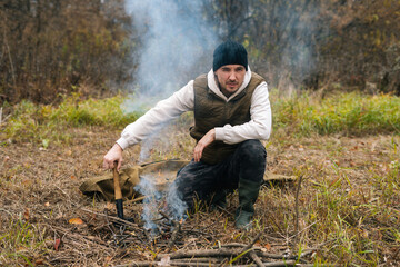 Portrait of confident tourist male wearing warm clothes sitting near burning campfire and holding small shovel, looking at camera. Concept of scout, research, travel and survival in nature. - Powered by Adobe