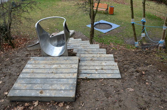 Stainless Steel Slide On The Playground With Concrete Hills And Rocks And Wooden Staircase. Large Glossy Tube For Sliding Children With An Impact On The Tartan Rubber Pad. The Tube Is An Open Trough