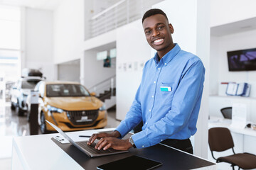 Cheerful black car salesman posing at work desk, smiling at camera, typing on laptop in auto...