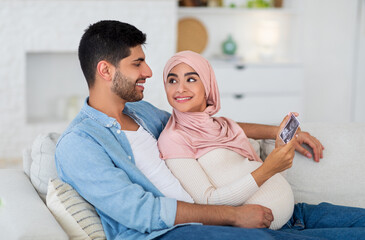 Obraz premium Happy islamic spouses looking at ultrasound photo of their future baby resting at home on sofa, free space