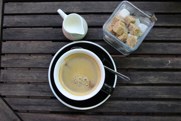 front top photo of a cup of freshly brewed coffee on a brown wooden table with a milk pot and sugar bowl 