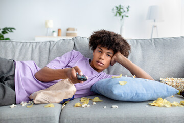 Lazy black teen guy lying on couch with scattered snacks, holding remote control, watching TV at...