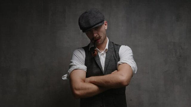 Portrait of young handsome confident rude mustachioed man in vintage suit, cap looking at camera, kneads his hands and standing arms crossed on concrete background. Mens power promotion theme template