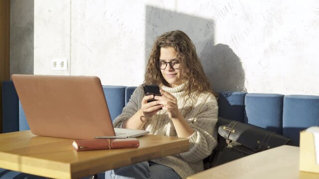 Pretty long haired businesswoman in warm sweater and glasses with smartphone in hands working on contemporary laptop at small table near large window in sunny cafe
