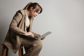 Young woman with poor posture using laptop while sitting on stool against grey background, space for text - obrazy, fototapety, plakaty