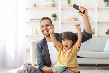 Positive father watching football game on tv with his little son, emotional boy shouting enjoying...