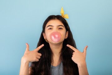 Funny Indian teen girl blowing bubble from sugar free chewing gum, pointing at it over blue studio background