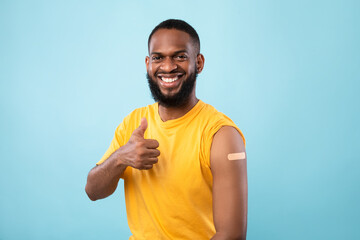 Cheerful Afro man showing thumb up gesture, demonstrating arm with band aid after injection of coronavirus vaccine