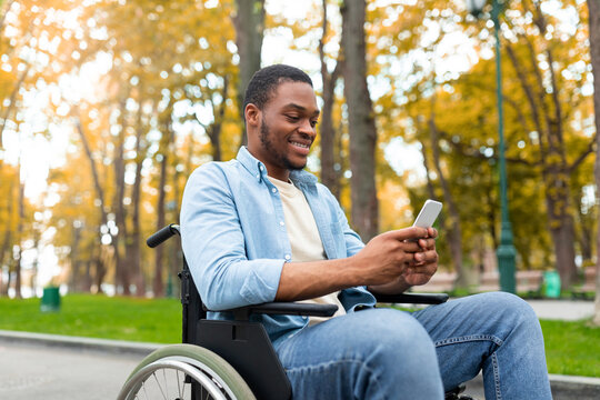 Cheery disabled black guy in wheelchair using mobile phone, checking email or messages at autumn park