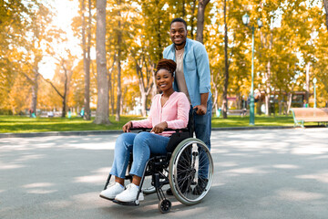 Full length of cheerful black guy taking his impaired girlfriend in wheelchair for walk at city park in autumn