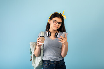 Indian teen female student with backpack and headphones holding takeaway coffee, using smartphone on blue background