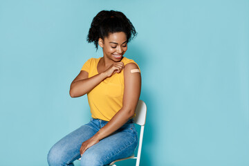 Smiling black woman getting vaccinated against covid, showing arm with band aid on blue background, free space - obrazy, fototapety, plakaty