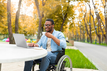Remote job for impaired people. Cheerful black guy in wheelchair working online, using laptop at outdoor cafe in autumn