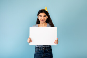 Amazing promo. Happy Indian teen girl holding blank white banner with copy space on blue studio...
