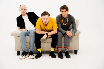 Young Guys Sit on a Sofa on a White Background and Look Together at the Camera. Three Friends are Sitting on the Couch and Watching TV, Watching a Home Theater Together. Close-up