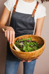 Woman in a gray apron mixing green salad in eco wooden bamboo bowl in the kitchen. Healthy eating and nutrition, eco conscious lifestyle.