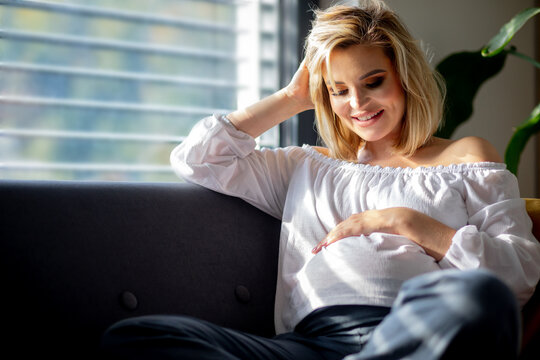 Pregnant rest at cozy home woman sitting on sofa and smiling to her belly