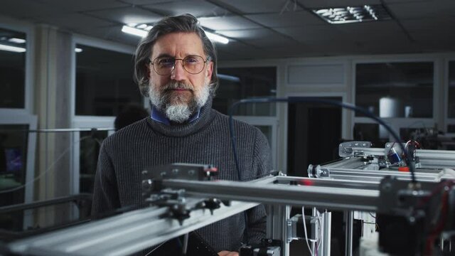 Bearded middle aged male engineer in turtleneck and glasses looking at camera while standing behind modern 3D printer printing model in professional studio