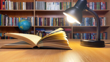 Book on wooden table under lamp light in classic library. Wind turning pages. Bookshelves.