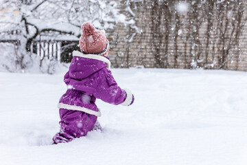 Little girl in violet winter overall, brown hat and violet gloves walking in deep snow in the garden. Small girl stepping into knee deep snow during snowfall. Young child walks in snow at winter