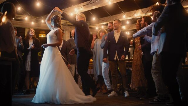 Beautiful Bride in White Dress and Groom in Stylish Black Suit Celebrate Wedding at an Evening Reception Party. Newlyweds Dancing at a Venue with Best Multiethnic Diverse Friends.
