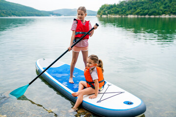 Girls padding on stand up paddle boarding on lake district. Children in swim life vest learning paddleboarding on SUP board. Family active leisure and local getaway with kids concept - obrazy, fototapety, plakaty