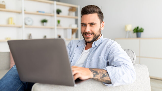 Positive guy reclining on sofa with laptop at home interior, urfing internet or scrolling social networks, panorama