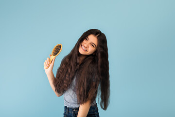 Attractive Indian teenage girl with long silky dark hair holding wooden brush, posing over blue studio background