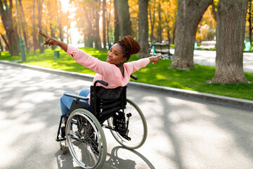 Young disabled black woman in wheelchair spreading arms, making victory gesture, feeling free at autumn park