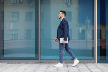 Fototapeta na wymiar Cheerful arab businessman with pad walking by street, full length
